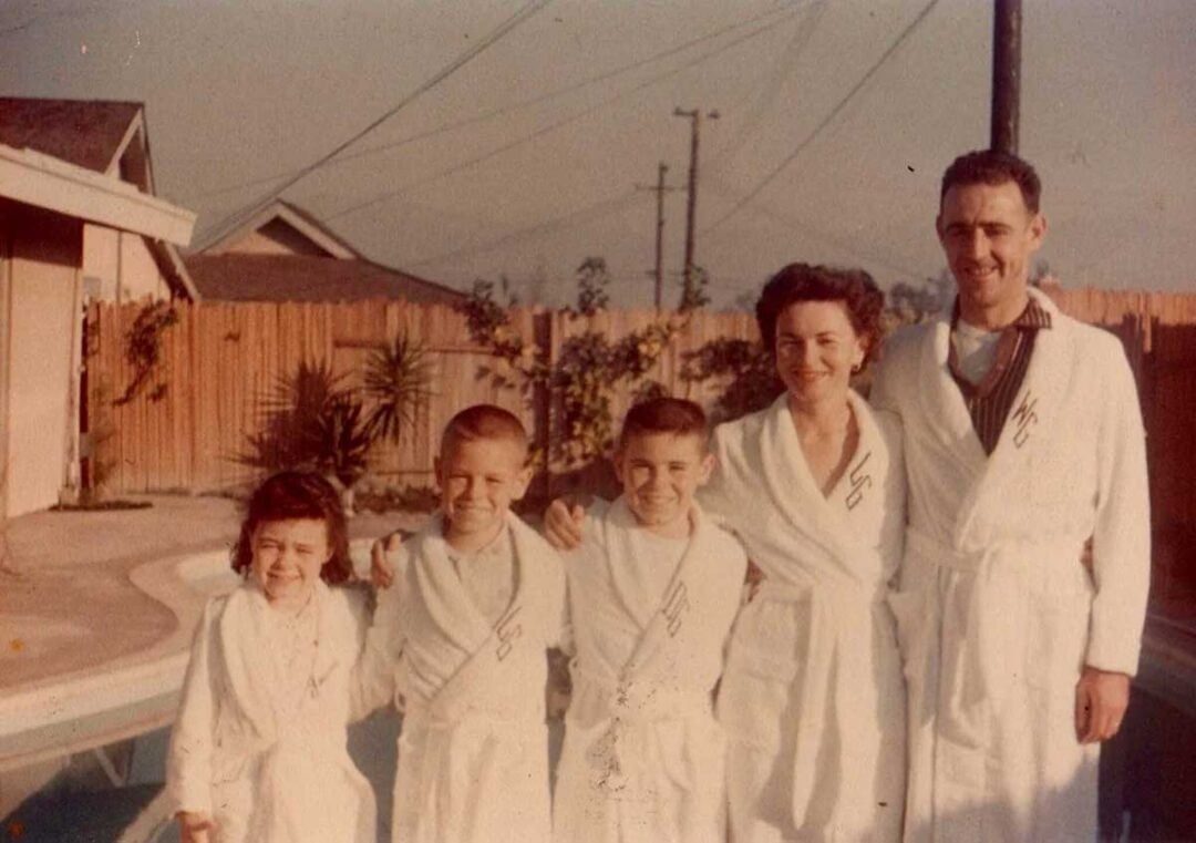 A family of five stands by a backyard pool, smiling and wearing matching white robes. Three children are between a woman and a man, and a wooden fence and houses are visible in the background.