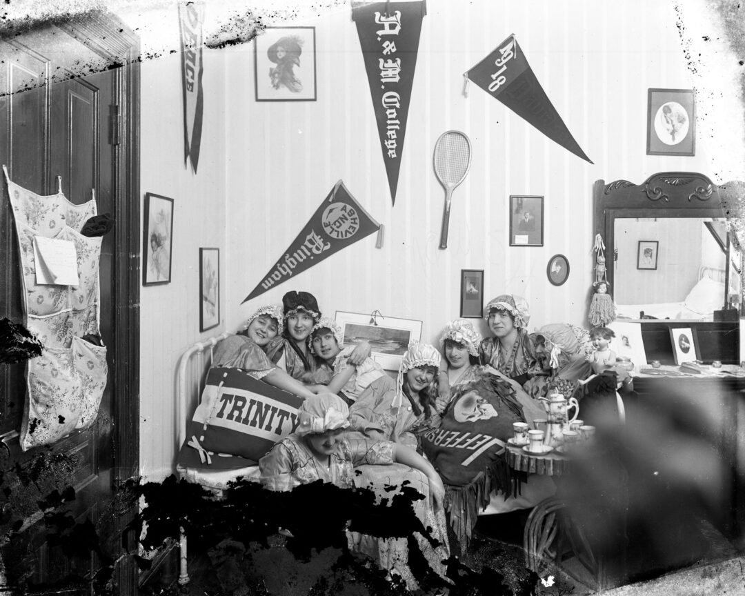 Five women in robes and bonnets sit on a bed in a decorated room with college pennants, drinking tea. The room has framed photos and a mirror. The photo is black and white and appears vintage.