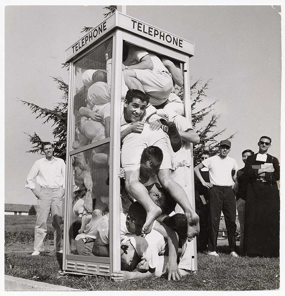 A group of people, mostly young men, are crammed into a small glass telephone booth, with some limbs and heads pressed against the glass, while others stand outside watching and smiling.