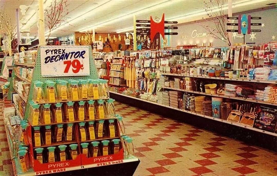 A vintage store aisle with red and white checkered floors, shelves stocked with various household goods, and a display of Pyrex decanters priced at 79 cents. Retro holiday decorations hang from the ceiling.