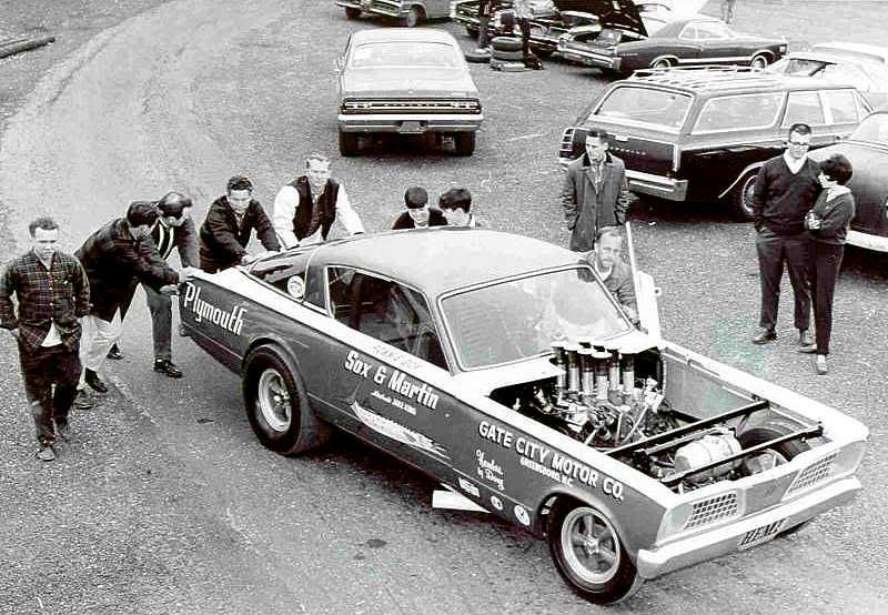 A group of people pushes a vintage Plymouth race car with exposed engine in a parking lot, surrounded by classic cars. The car features sponsor logos and "Sox & Martin" on the side.