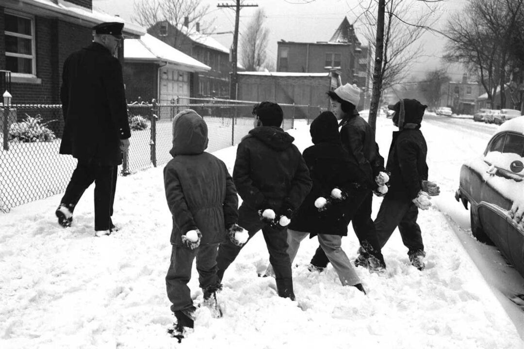 Six children holding snowballs stand in the snow, appearing ready to playfully throw them at a police officer walking ahead on a snowy residential street lined with houses and a parked car.