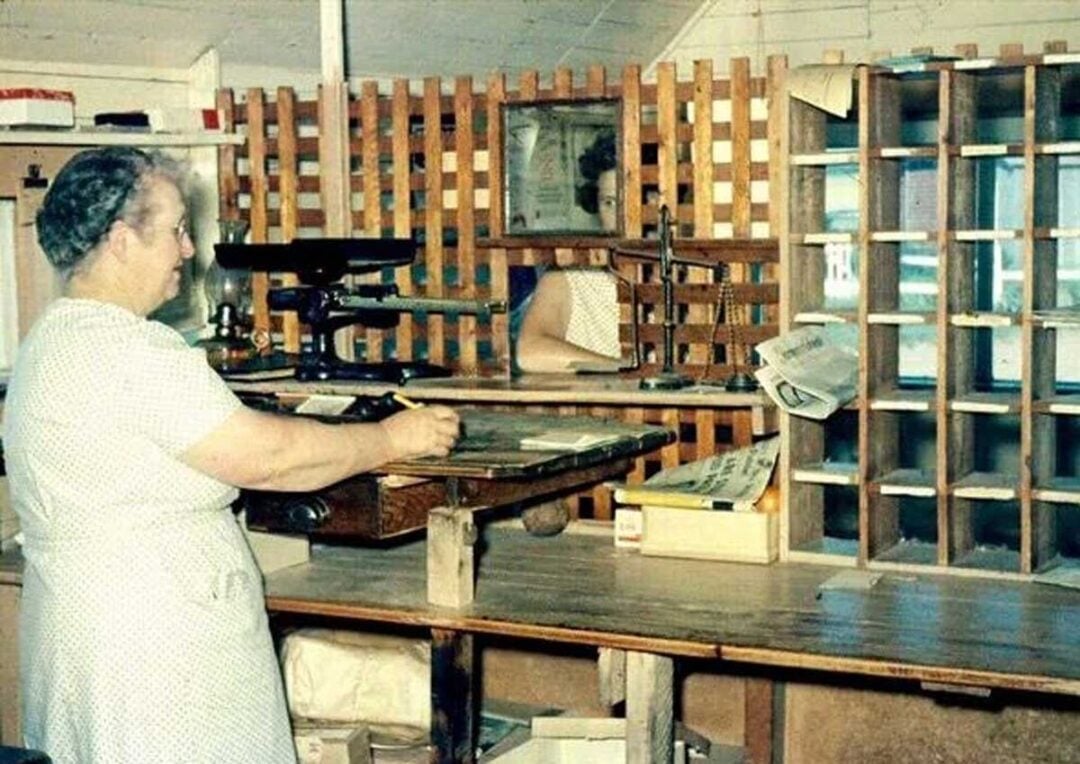 An older woman stands at a wooden counter in a vintage-style office or post office, sorting papers with cubbyholes and shelves in the background, and a mirror reflecting her image.