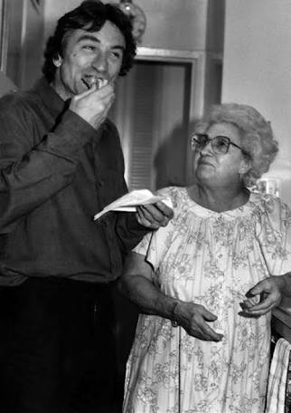 A man stands in a kitchen eating food from a napkin, smiling, while an older woman wearing glasses and a floral dress looks up at him with a gentle expression. The image is in black and white.