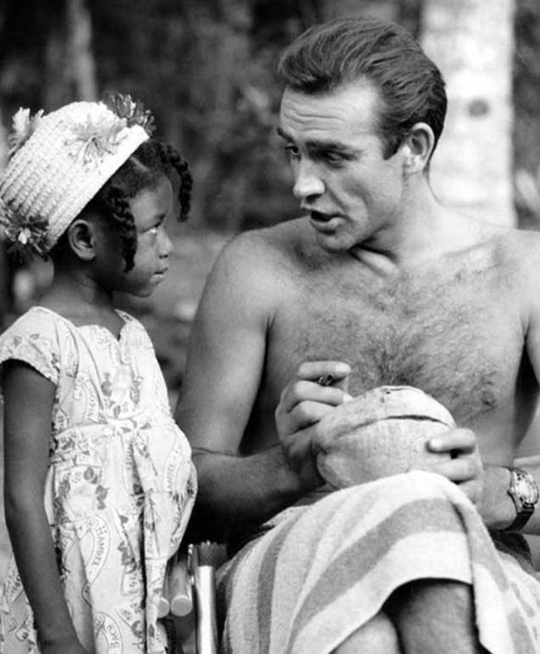 A shirtless man with a towel wrapped around his waist sits holding a coconut, talking to a young girl in a dress and hat with flowers. They are outdoors, and both appear engaged in conversation.
