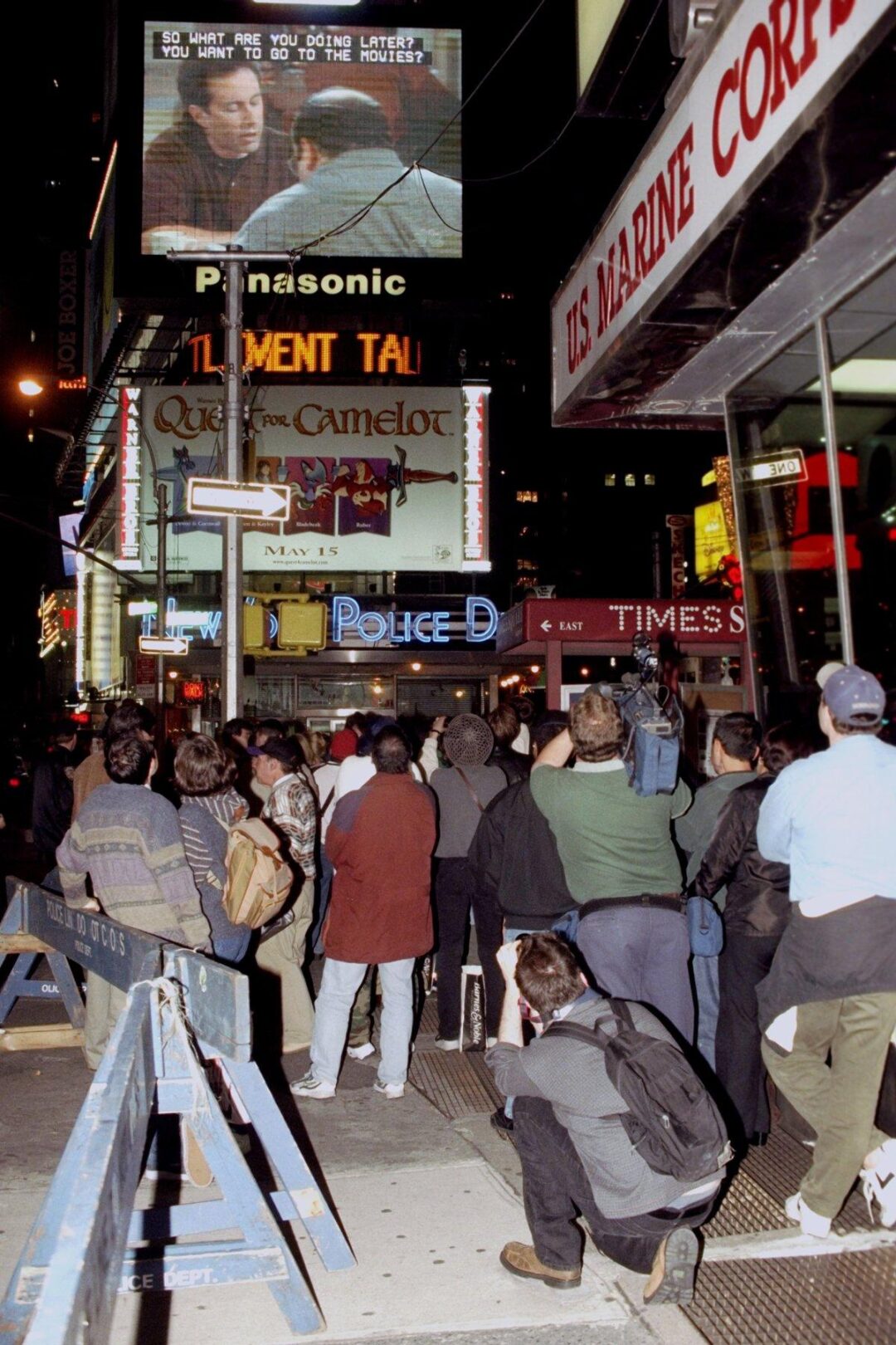 A crowd of people stands on a busy street at night, watching a large outdoor screen showing a movie scene with subtitles in Times Square, New York City, surrounded by bright signs and storefronts.