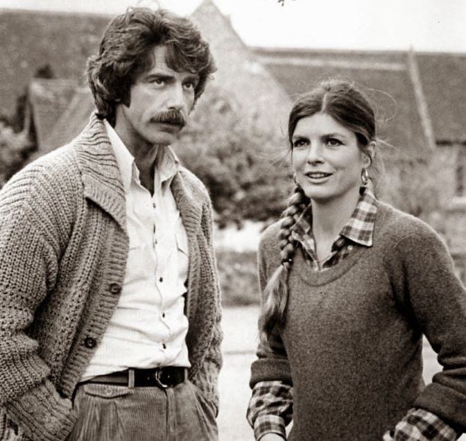 A black and white photo of a man with a mustache wearing a cardigan and button-down shirt, standing next to a woman with braided hair in a sweater and plaid shirt. They are outdoors with buildings in the background.