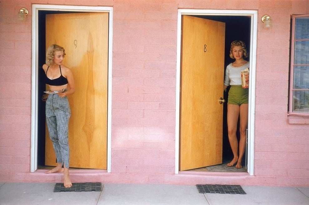 Two women stand barefoot in adjacent doorways of a pink building. One, in high-waisted pants and a black top, holds a plate; the other, in green shorts and a white shirt, holds a cereal box. Both doors are wooden, numbered 8 and 9.