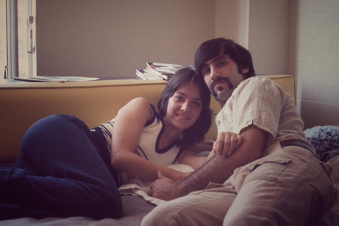 A smiling woman and man lie side by side on a bed, leaning against each other in a cozy room with sunlight coming through a window and a stack of papers in the background.