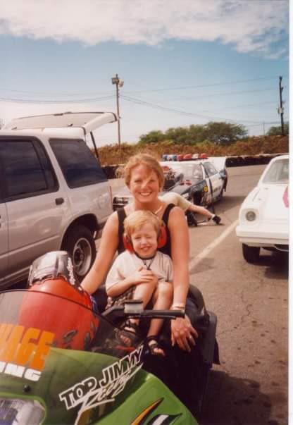 A smiling woman and a young child sit together on a green jet ski in a parking lot, surrounded by cars under a partly cloudy sky.