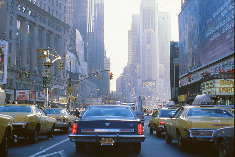A busy city street filled with yellow taxis and cars, surrounded by tall buildings and billboards under a hazy sky, capturing the hustle of urban life.