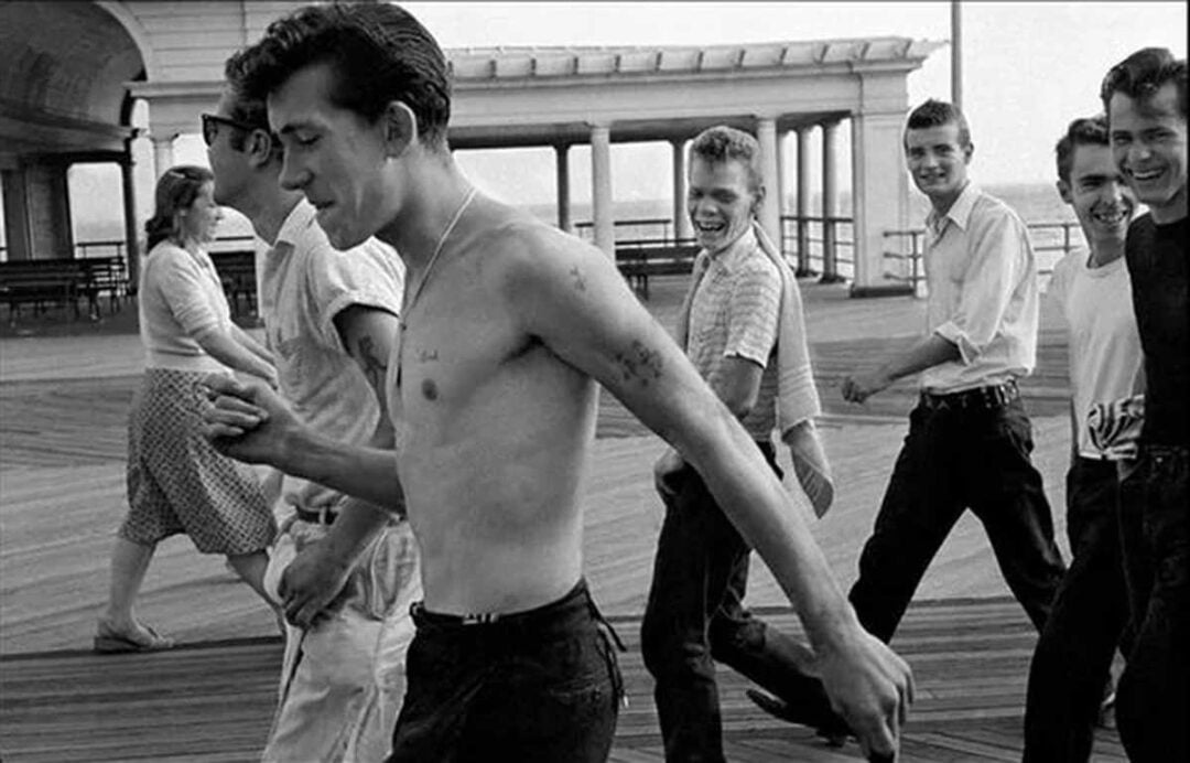 A group of young men, one shirtless, walk and smile on a wooden boardwalk by a pavilion, with a few people in the background. The scene appears lively and candid, with a sense of carefree youth.