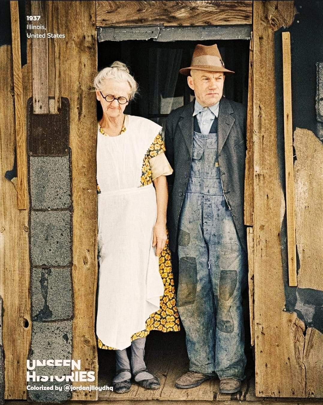 An elderly woman in a white apron with yellow flowers and a man in overalls, coat, and fedora stand in a rustic wooden doorway. The photo is colorized and labeled “1937 Illinois, United States.”
