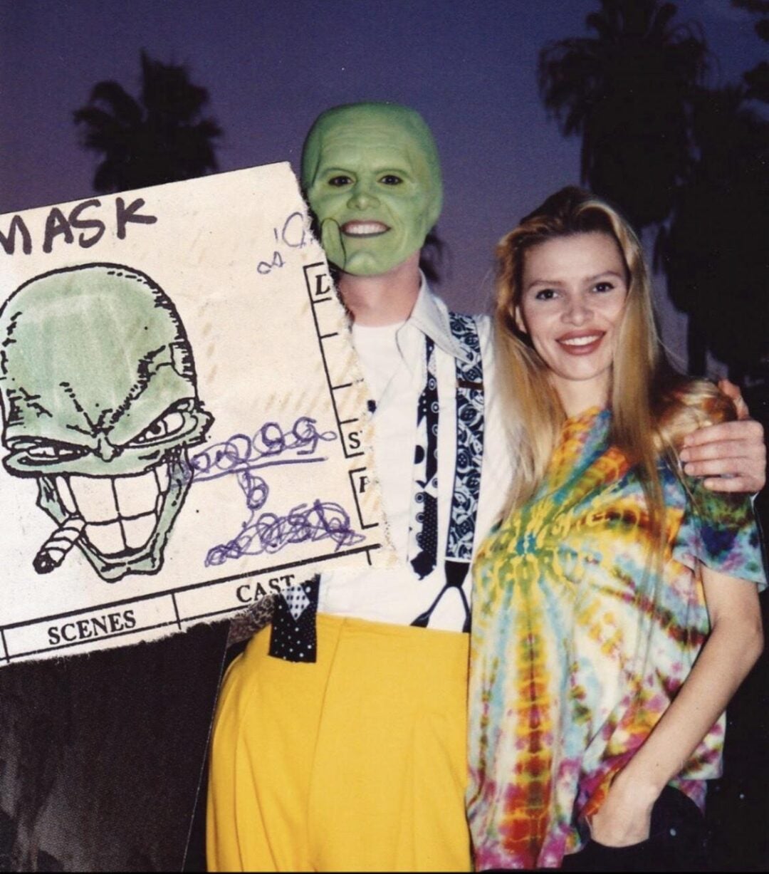 Two people pose outdoors. One wears a bright green mask, yellow pants, a patterned tie, and holds a large “The Mask” poster. The other person, a woman, smiles and wears a colorful tie-dye shirt. Palm trees are visible in the background.