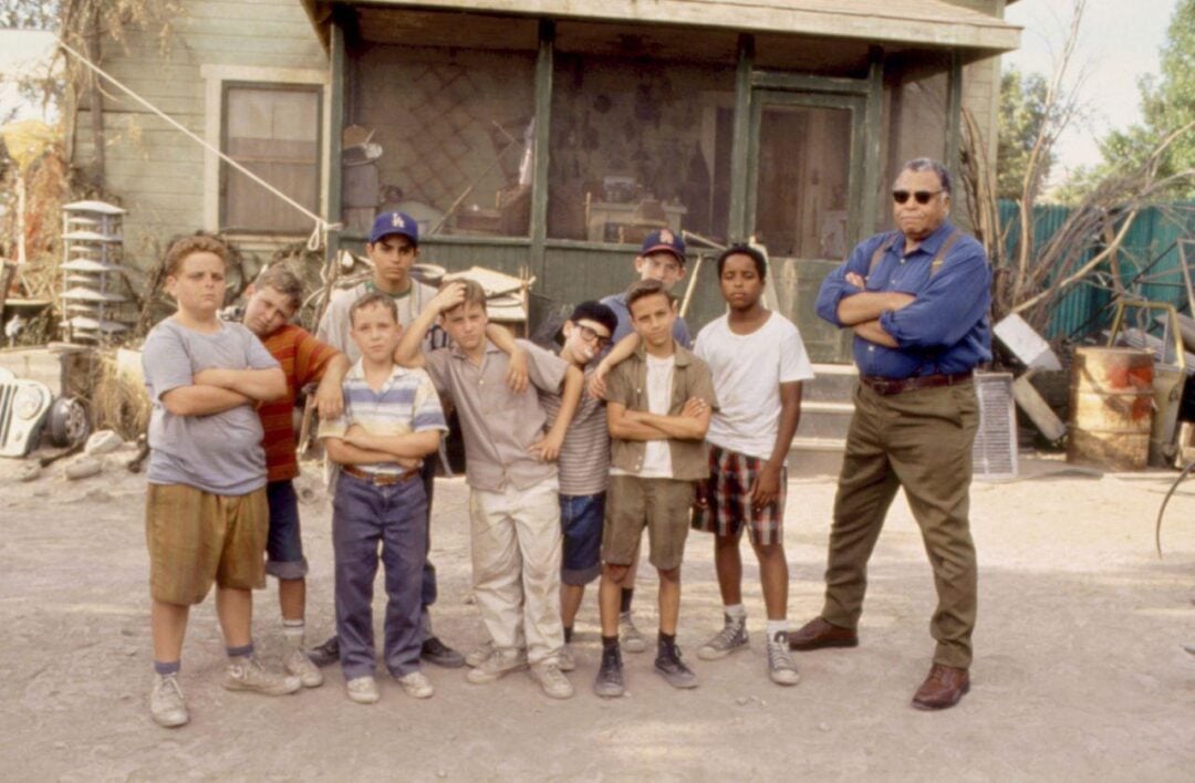 A group of nine boys stand with their arms crossed next to a tall man in sunglasses, also with arms crossed, in front of a rundown house. The boys wear casual clothes and caps, looking serious.