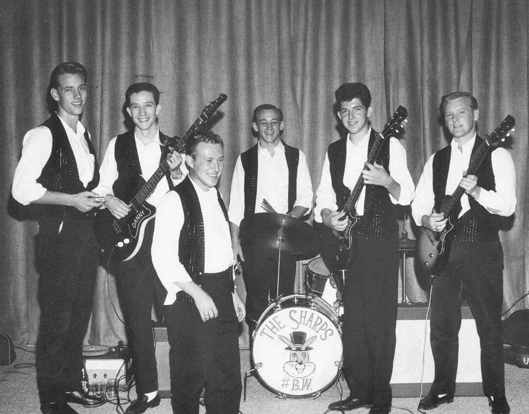 Six young men in matching vests and white shirts pose with musical instruments on stage. Five hold guitars and one sits at a drum set labeled “The Sharps #B.W.” A curtain hangs behind them.