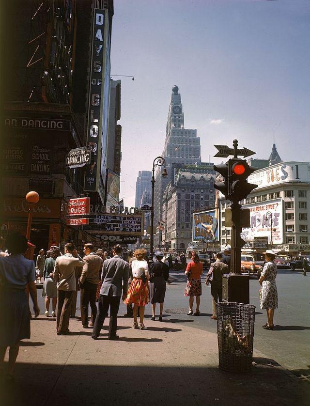 People stand at a crosswalk in a busy city street, surrounded by tall buildings with vintage signs and billboards. A red traffic light hangs overhead, and cars are visible in the background. The scene appears to be from mid-20th century.