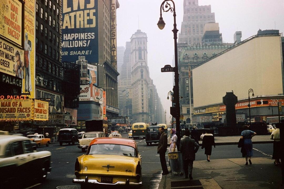 A busy city street scene in Times Square, New York City, with vintage cars, people walking and billboards advertising theaters and shows. The image appears to be from the mid-20th century.