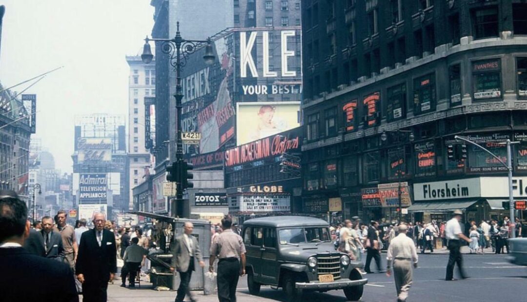 A busy city street scene from the mid-20th century with crowds of people, classic cars, and vintage storefronts and signs, including “Klein’s,” “Admiral Television Appliances,” and “Florsheim.”