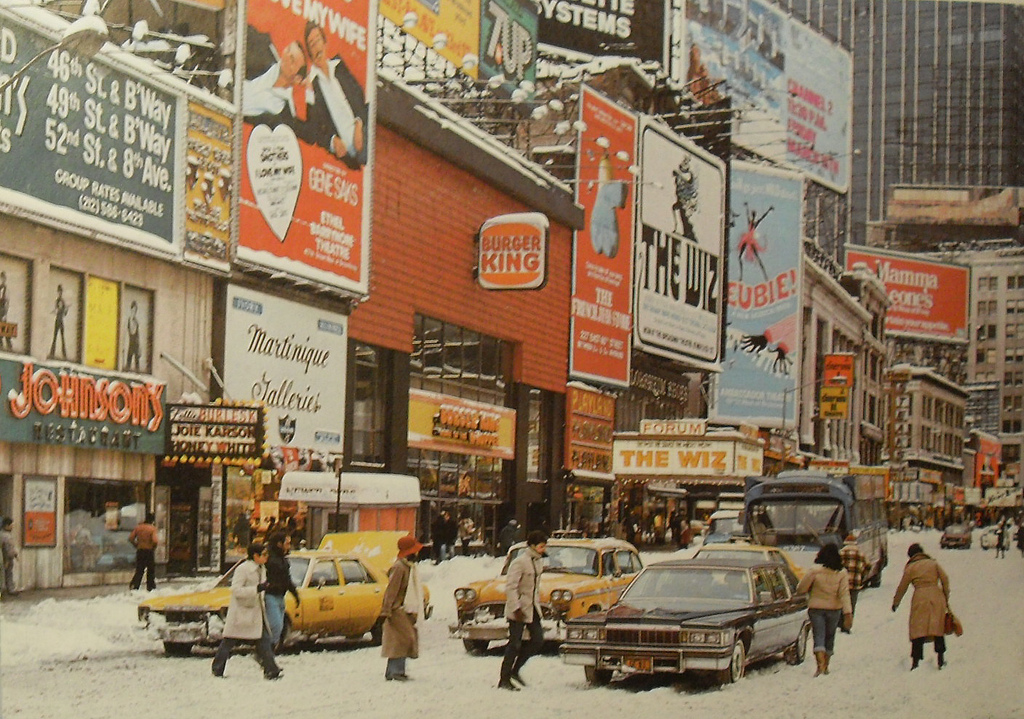 A busy city street in winter with people crossing, snow on the ground, yellow taxis, cars, and many colorful billboards, including signs for Burger King and The Wiz. Tall buildings surround the scene.