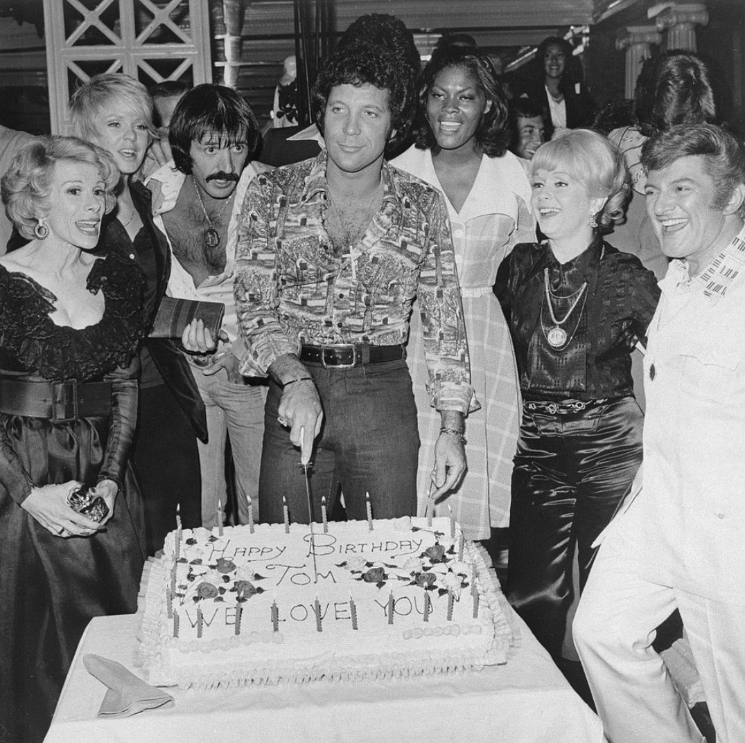 A group of people gathered around a man cutting a large birthday cake that says "Happy Birthday Tom, We Love You!" Everyone is smiling and dressed in formal or festive attire at what appears to be a party.