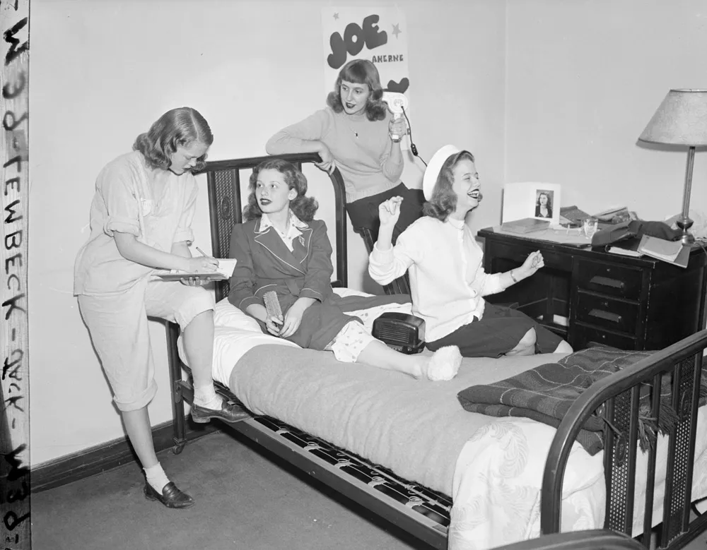 Four young women are gathered in a dorm room, smiling and talking. Three sit on a bed while one stands nearby holding a notepad. The room has a lamp, framed photo, and a "Joe" poster on the wall.