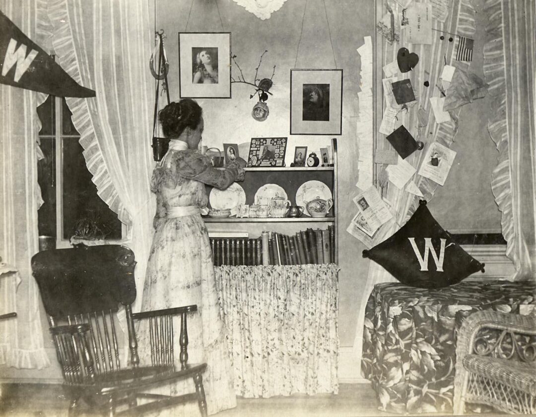 A woman in a long dress arranges items on a small shelf in a vintage room decorated with framed photos, books, a chair, and pennants with a “W.” Letters and papers hang from string by the bed, which has a floral bedspread.
