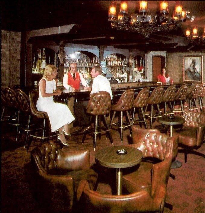 A vintage bar scene with tufted leather chairs, a chandelier, and a bartender serving a man and woman at the counter; another woman sits alone at a side table in the dimly lit, elegant room.