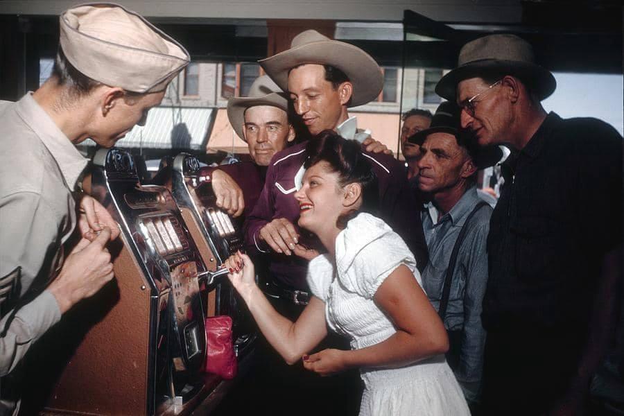A woman in a white dress smiles while pulling the lever on a slot machine, surrounded by men in hats and western attire, all watching her in a lively indoor setting.