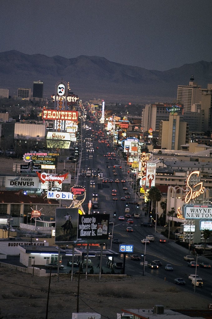 A view of a brightly lit Las Vegas street at dusk, lined with neon signs and billboards for casinos, hotels, and shows, with mountains visible in the background.