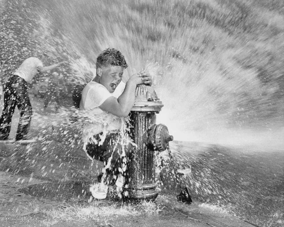 A boy in a white shirt smiles and clings to an open fire hydrant spraying water, while another child plays in the spray behind him, creating a joyful scene on a city street.