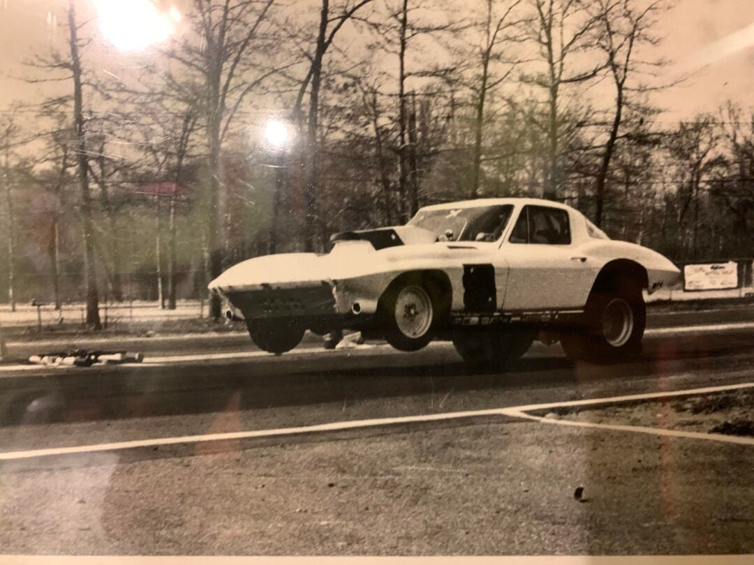 Black and white photo of a classic race car with its front wheels lifted off the ground, speeding down a track. Leafless trees and roadside signs line the background.
