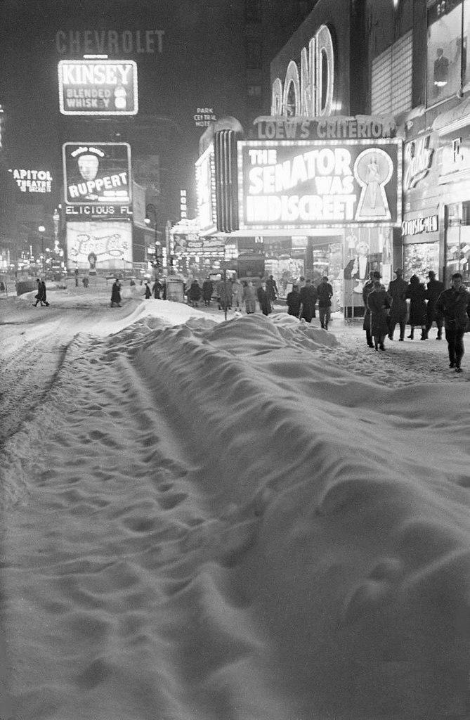 A snowy city street at night in the mid-20th century, lined with illuminated theater and advertising signs. People walk along sidewalks beside deep snowbanks under the bright lights.