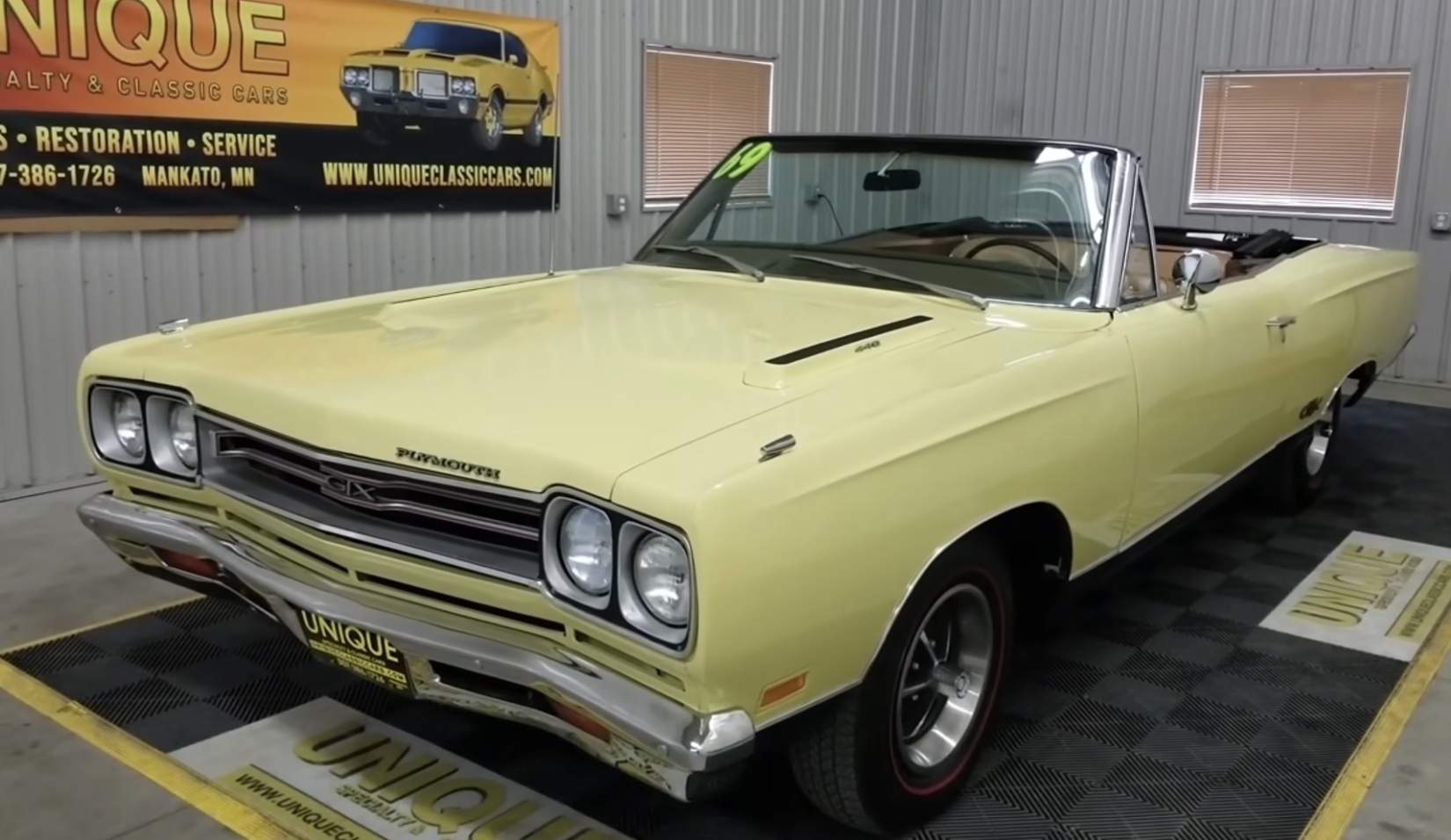 A yellow classic Plymouth convertible car with a black interior is displayed indoors on a black mat. The car has its top down and is parked in front of a sign for Unique Classic Cars.
