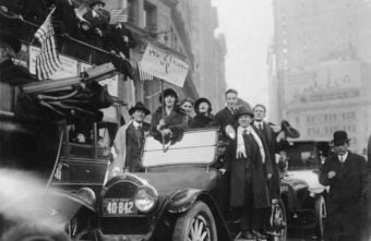 A group of people, both men and women, stand and sit on an old-fashioned car in a city street, waving flags and holding a sign that reads &ldquo;We Cleaned Up!&rdquo; during a lively parade or celebration.