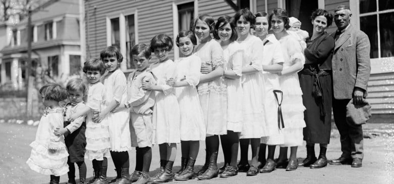 A group of twelve girls in white dresses stand in a line outdoors, arms linked. Two small children stand in front, while a smiling woman holding a baby and a man stand together at the end of the line near a house.