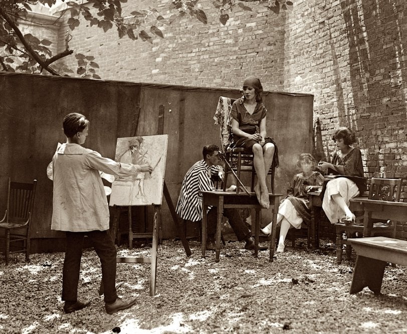 A group of people are outdoors; one person paints at an easel while a woman poses on a chair. Three others sit nearby, watching or drawing, surrounded by chairs and a brick wall under tree shade.