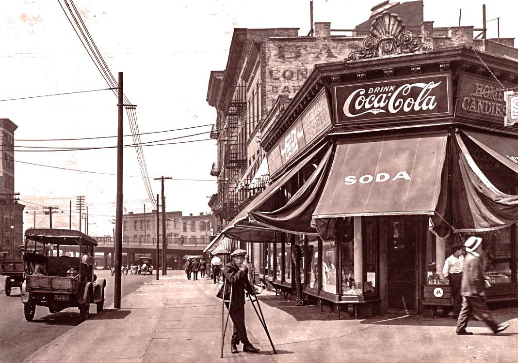Historic city street scene with people walking, a vintage car parked, and a corner store featuring a large Coca-Cola sign and "SODA" awning. A man stands with a camera tripod on the sidewalk. Early 20th-century setting.