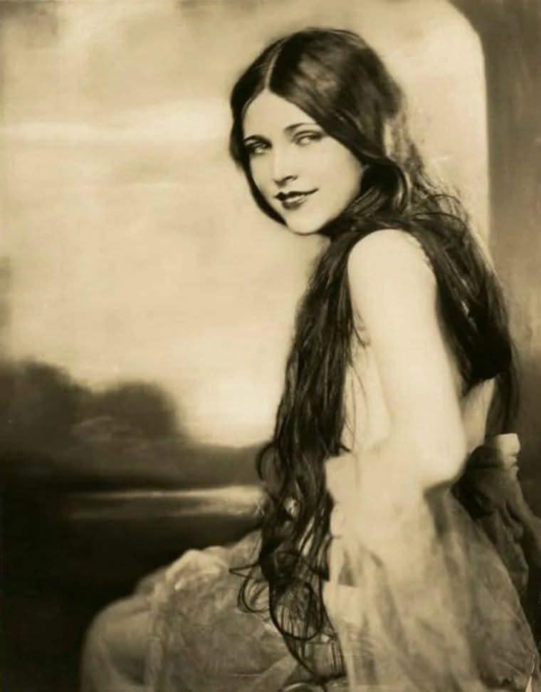 A vintage black-and-white portrait of a woman with long, dark hair, wearing a sleeveless dress and looking toward the camera with a gentle smile. The background is softly blurred, giving the image an ethereal feel.