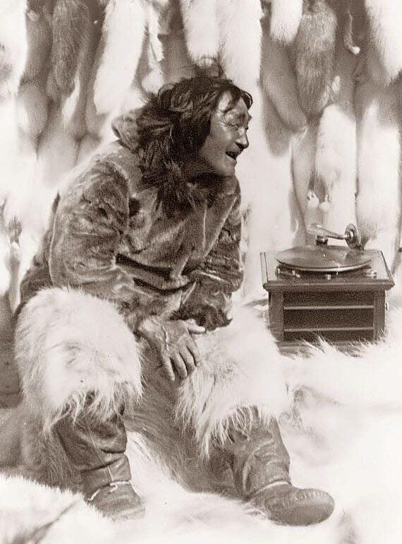 A person wearing traditional fur clothing sits on fur pelts, smiling and listening to a vintage gramophone. The background is lined with more pelts.