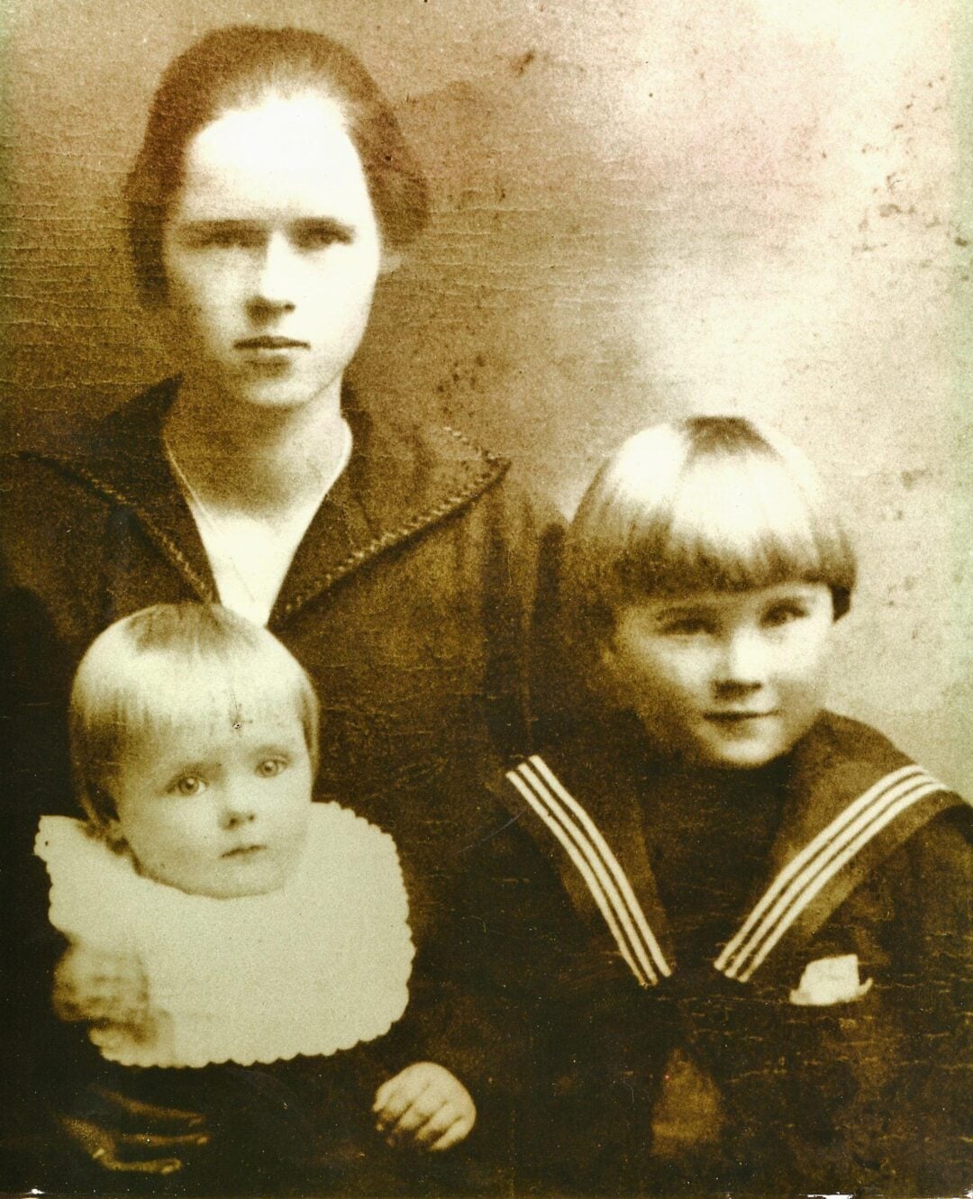 Sepia-toned vintage portrait of a woman with short hair sitting beside two young children, one a baby in a white gown and the other an older child in a sailor outfit, all looking at the camera.