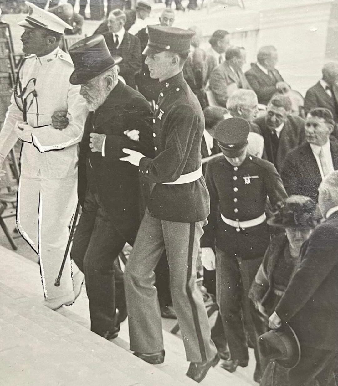 An elderly man with a cane is helped up steps by uniformed officers. A crowd of men and women, some in uniform, follow behind, suggesting a formal or official event. The image is black and white.