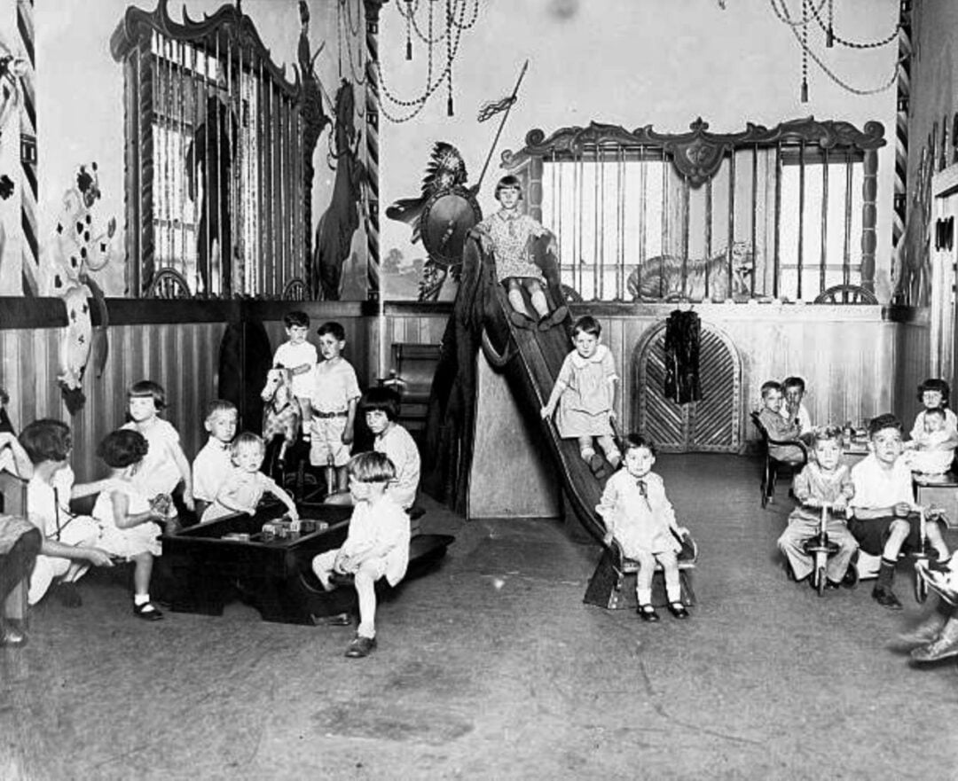 Black-and-white photo of young children playing in a decorated indoor room with a slide, small tables, and toys. The walls feature murals of animals and circus-themed decorations. An adult stands by the slide, watching the children.