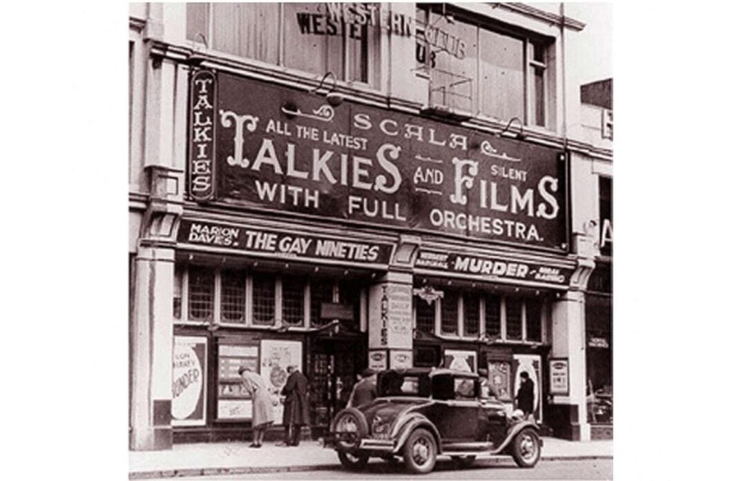 A vintage photograph of a theater showing films, with a marquee advertising “Talkies and Silent Films with Full Orchestra.” People stand near the entrance and a classic car is parked in front.