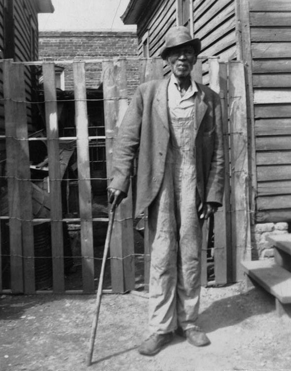 An elderly man wearing a hat, coat, and striped overalls stands outside between wooden buildings, holding a cane and facing the camera. A wooden fence and steps are visible in the background.