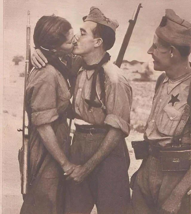 A woman and a man in military uniforms share a kiss while another soldier stands beside them, smiling. They appear to be outdoors, likely during a historical wartime setting.