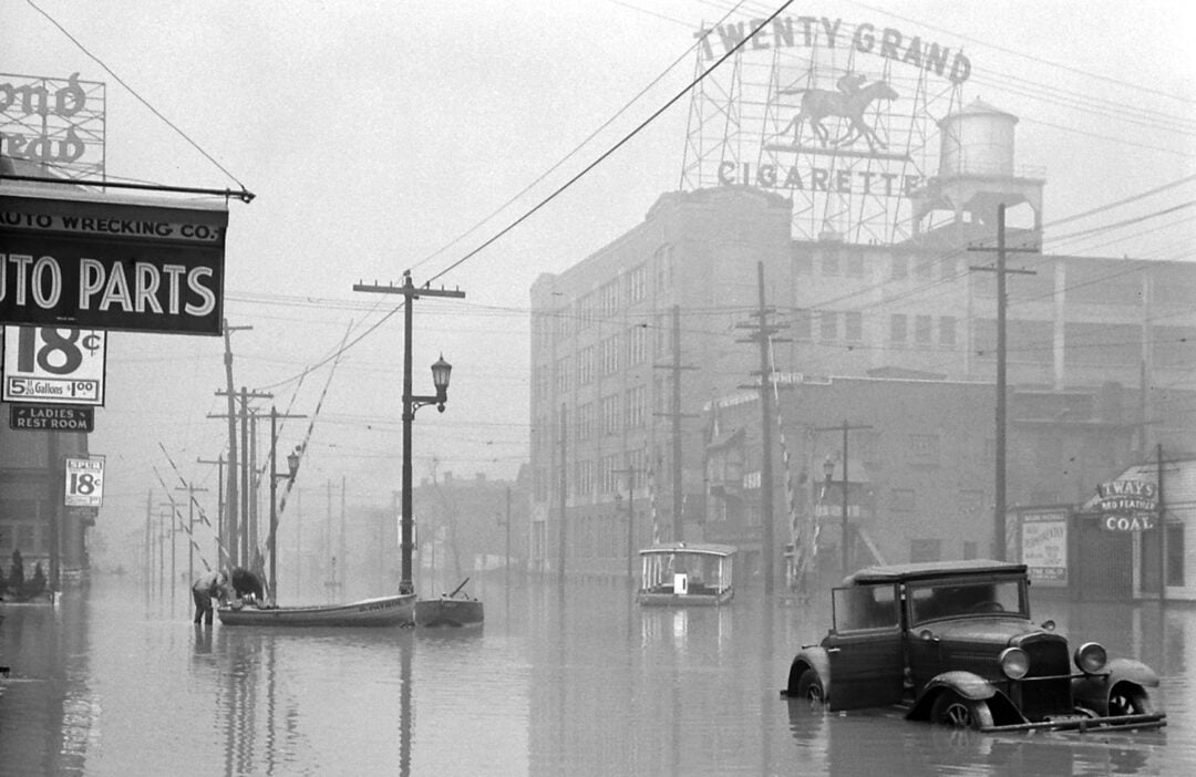 A black-and-white photo shows a city street flooded with water; a stranded car and a boat are visible, along with power lines, old buildings, and a large "Twenty Grand Cigarettes" sign in the background.
