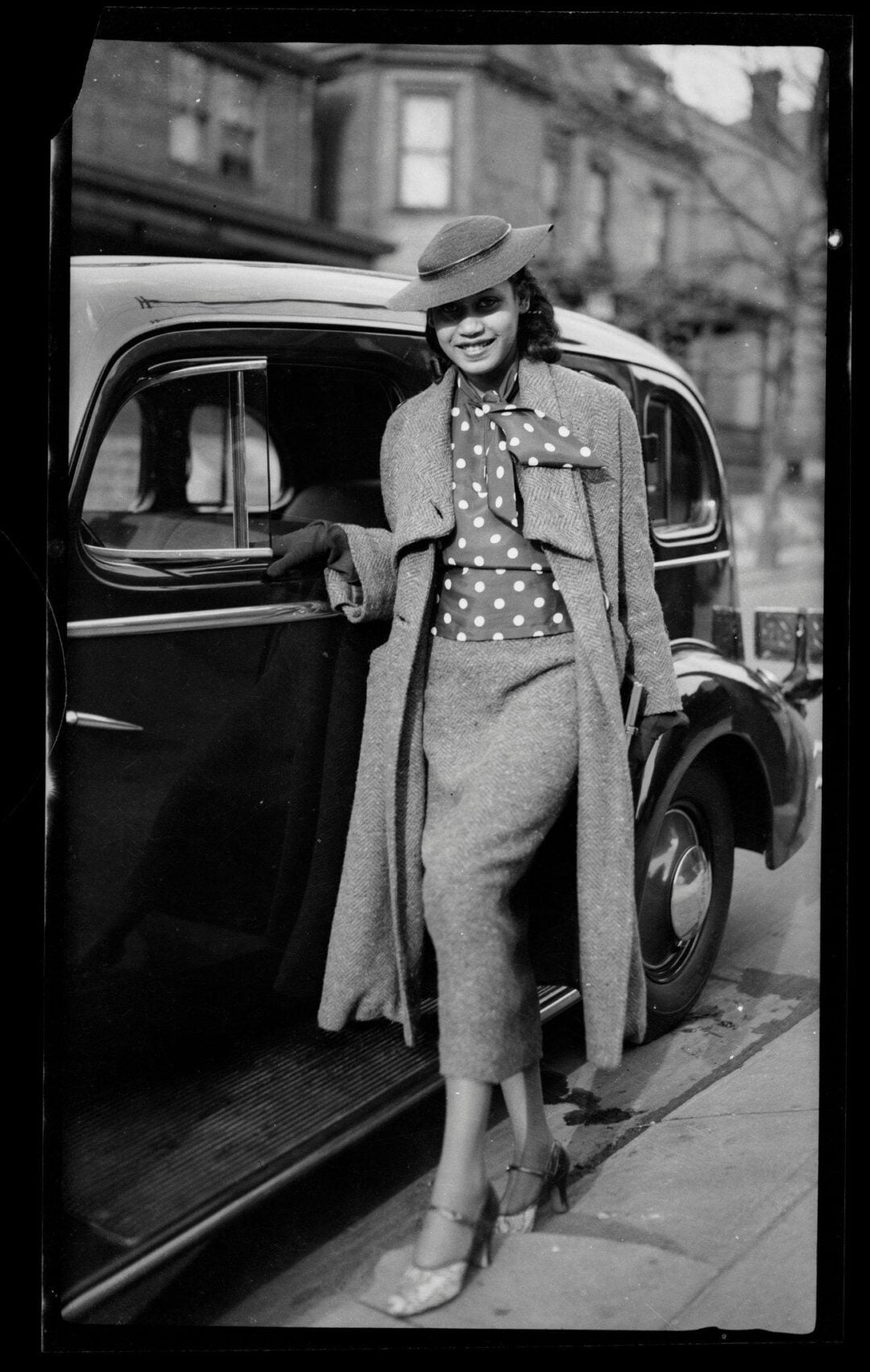 A stylish woman in a long coat, polka-dot blouse, and hat smiles while leaning against a vintage car on a city street. The photo appears to be from the early to mid-20th century.