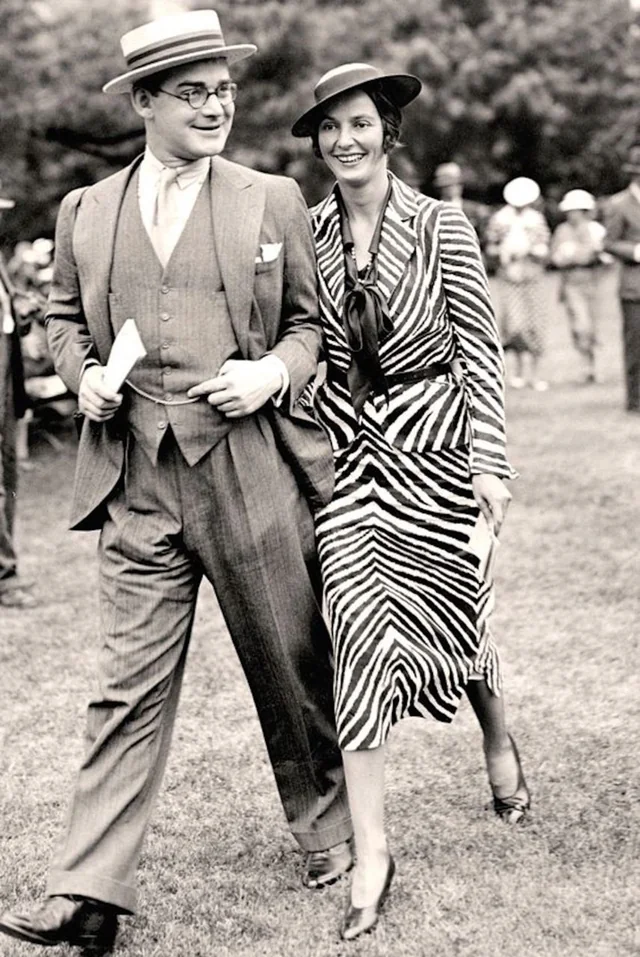 A man in a suit and straw hat walks beside a smiling woman wearing a striped dress, hat, and heels. They appear to be at an outdoor event, with other people and trees visible in the background.
