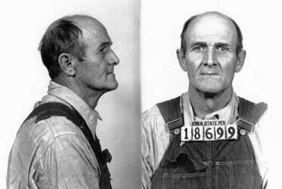 Black and white mugshot of an older man with thinning hair, wearing overalls and a collared shirt. He faces left in the side view and forward in the front view, holding a placard labeled "186999.